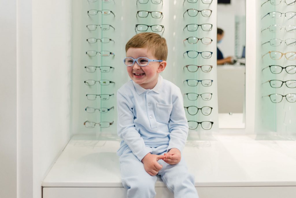 A smiling young boy, wearing glasses and sat on a counter in front of our eyewear range